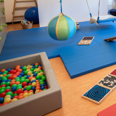 Ein Raum für Psychomotorik zeigt bunte Bälle, eine Schaukel und Texturmatten für spielerische Bewegungstherapie.