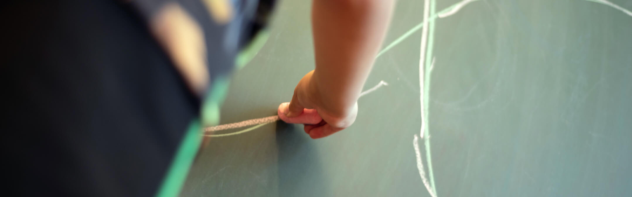 Ein Kind zeichnet mit Kreide Kreise auf eine grüne Tafel, während es sich über die Oberfläche lehnt.