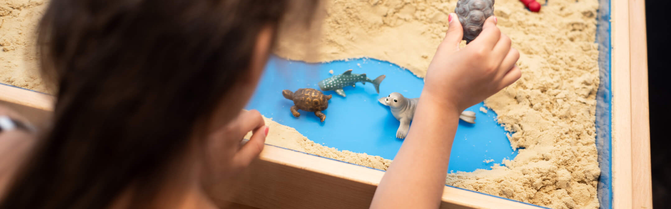 Ein Kind spielt mit Tierfiguren im Sandkasten und stellt fantasievolle Szenen am Strand nach.