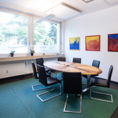 Ein heller Beratungsraum mit rundem Holztisch, modernen Stühlen und bunter Kunst an der Wand bietet eine einladende Atmosphäre.