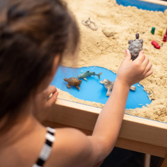 Ein Kind spielt mit Tierfiguren im Sandkasten und stellt fantasievolle Szenen am Strand nach.