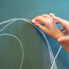 Eine Hand hilft einem Kind, mit Kreide auf einer Tafel zu zeichnen, was auf eine unterstützende Lerntherapie hinweist.