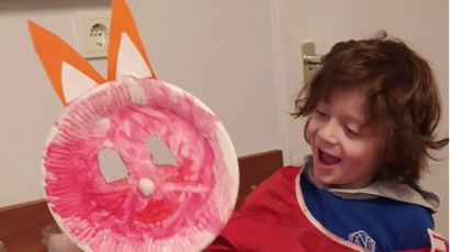 Ein lachendes Kind hält eine bunte Osterhasenmaske aus einem Pappteller mit rosa Farbe und orangefarbenen Ohren.