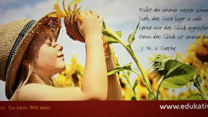 Ein Mädchen mit Strohhut riecht an einer Sonnenblume im Sonnenlicht, begleitet von einem Zitat von Goethe.