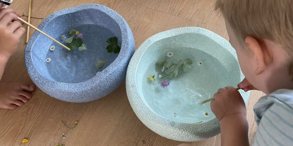 Zwei Kinder spielen im Sommer mit Blumen in Wasserschalen, wobei sie Stöcke benutzen, um die Blüten im Wasser zu bewegen.
