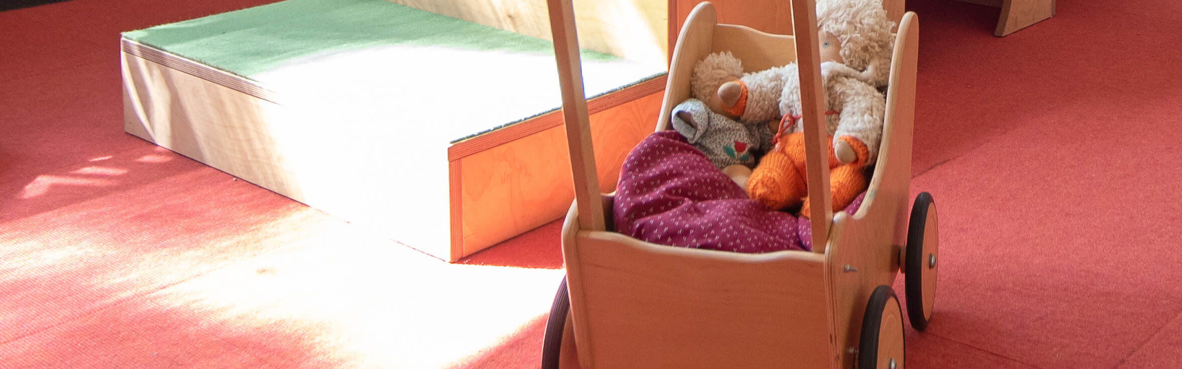 Ein Holzspielzeugwagen mit Kuscheltieren steht auf einem roten Teppich vor einer kleinen Holzrampe, ideal für Spieltherapie.