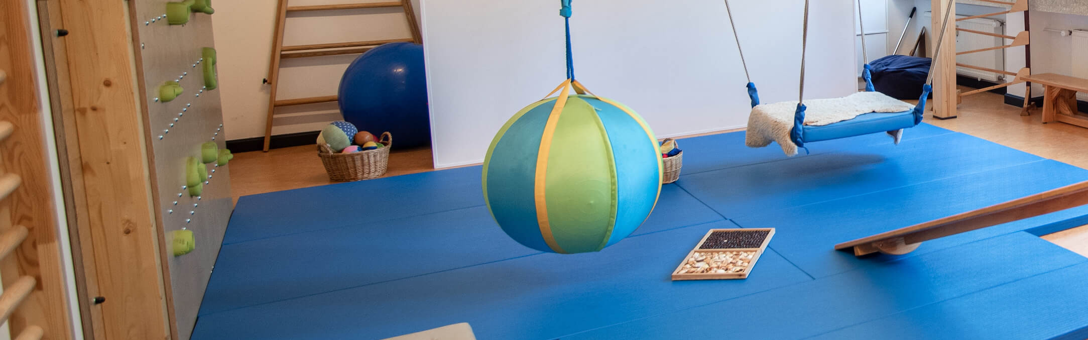 Ein Raum für Psychomotorik zeigt bunte Bälle, eine Schaukel und Texturmatten für spielerische Bewegungstherapie.