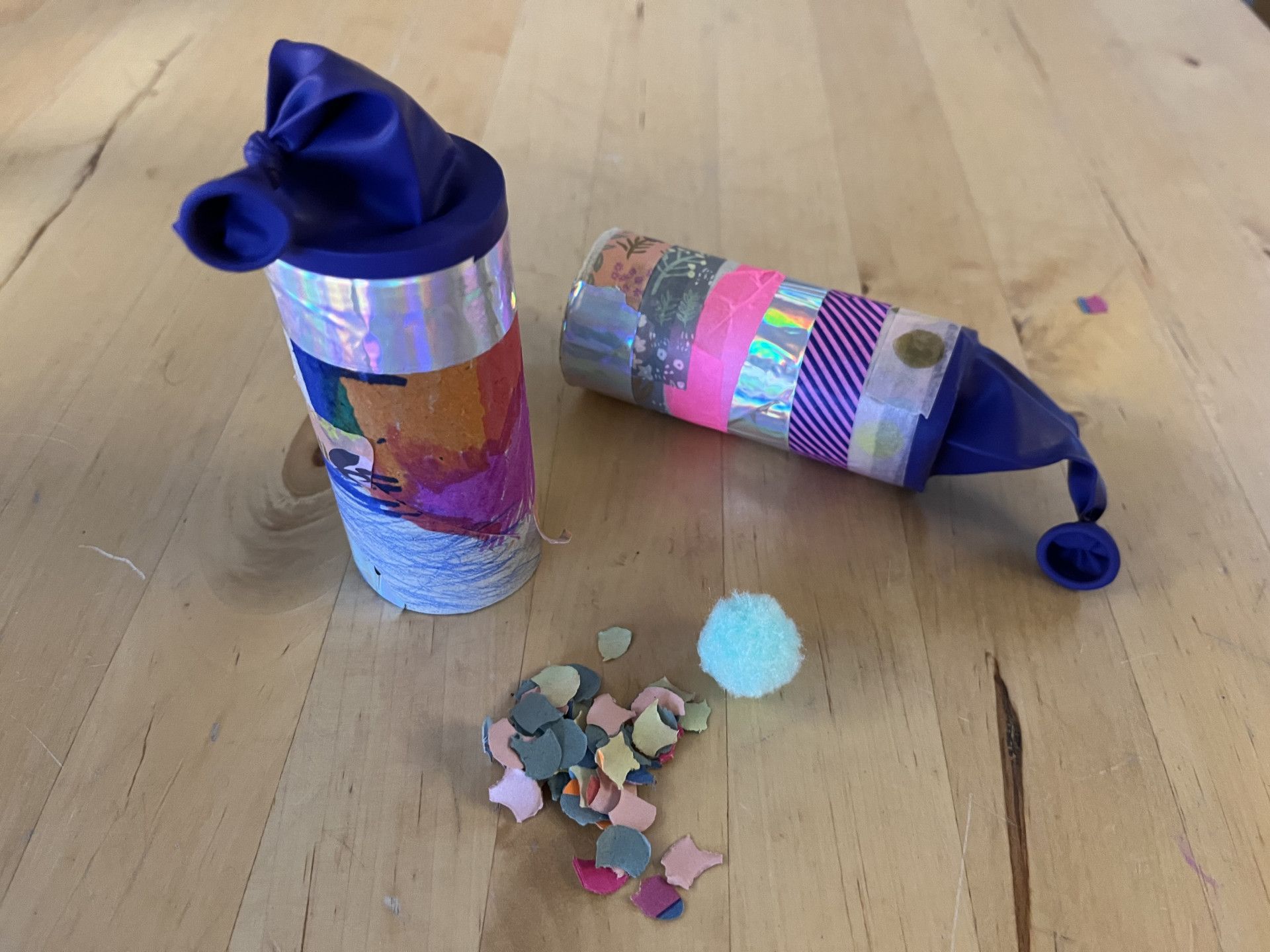 Zwei bunte, selbstgemachte Konfetti-Kanonen aus Papprollen und Luftballons liegen auf einem Holztisch mit Konfetti.