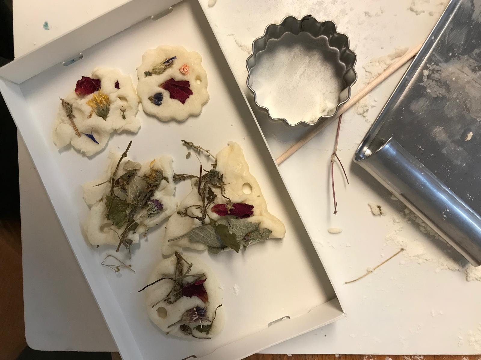 Salzteigformen mit getrockneten Blumen liegen auf einem Tisch neben einem Ausstecher und einem Tablett.
